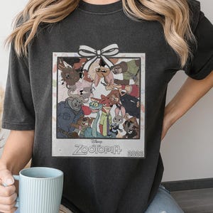 Puede incluir: Camiseta gris oscuro con un gr&aacute;fico enmarcado de personajes de la pel&iacute;cula de Disney Zootopia. El gr&aacute;fico incluye un lazo blanco y el texto "Zootopia 2026". La camiseta la sostiene una persona.