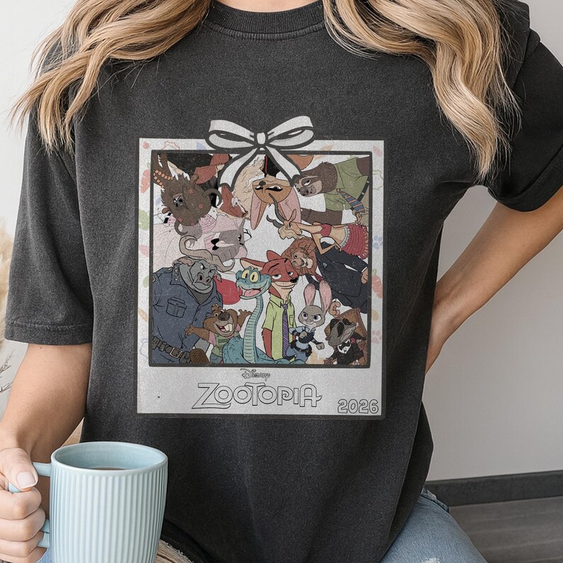 Puede incluir: Camiseta gris oscuro con un gr&aacute;fico enmarcado de personajes de la pel&iacute;cula de Disney Zootopia. El gr&aacute;fico incluye un lazo blanco y el texto "Zootopia 2026". La camiseta la sostiene una persona.
