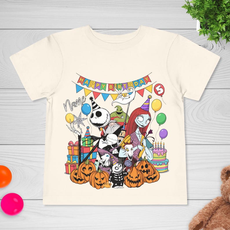 May include: Cream-colored t-shirt featuring a colorful graphic of characters from The Nightmare Before Christmas celebrating a birthday. The design includes pumpkins, presents, a cake, and the text "Happy Birthday". A number 3 is also visible.