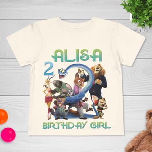 May include: Cream-colored t-shirt featuring a colorful graphic of animated animal characters and the name "ALISA" in green and blue. The number "2" and the words "BIRTHDAY GIRL" are also displayed. The shirt is likely for a child's birthday.