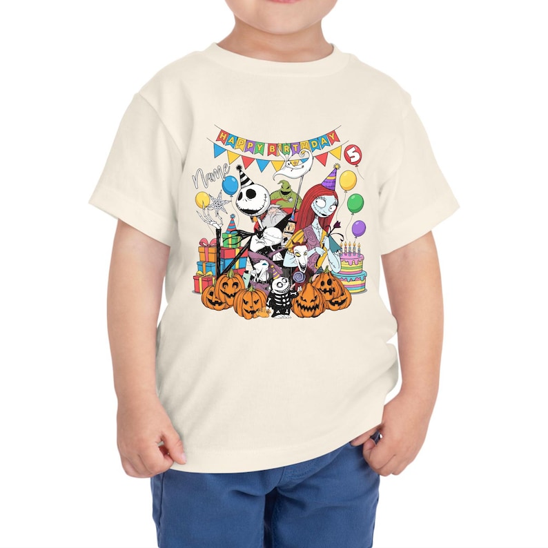 May include: A cream-colored toddler t-shirt featuring a colorful graphic of characters from The Nightmare Before Christmas. The design includes Jack Skellington, Sally, and pumpkins, with the text "Happy Birthday" and a number 5.