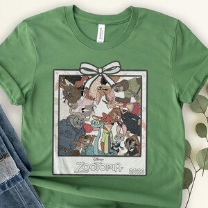 Puede incluir: Camiseta verde con una imagen enmarcada de personajes de la pel&iacute;cula de Disney Zootopia. La imagen tiene un lazo blanco y el texto "Zootopia 2026". La camiseta est&aacute; hecha de un material suave. Es una prenda informal.