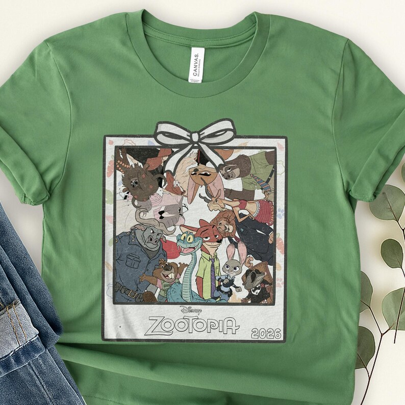 Puede incluir: Camiseta verde con una imagen enmarcada de personajes de la pel&iacute;cula de Disney Zootopia. La imagen tiene un lazo blanco y el texto "Zootopia 2026". La camiseta est&aacute; hecha de un material suave. Es una prenda informal.