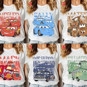 May include: White t-shirts with colorful graphics of characters from the animated movie Cars. The designs include Lightning McQueen, Sally, Mater, Mack, Doc Hudson, and Fillmore. Each shirt features a different character or group of characters.