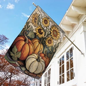 May include: A decorative outdoor flag featuring a fall harvest design. The flag displays a vibrant illustration of pumpkins in shades of orange and white, alongside sunflowers with yellow petals. The artwork is set against a backdrop of green foliage and a blue sky.