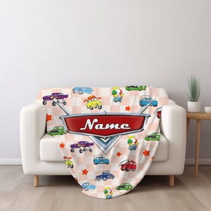May include: A soft blanket featuring a repeating pattern of cartoon cars in various colors on a pink and white checkered background. A large red emblem with the word "Name" in white script is prominently displayed.