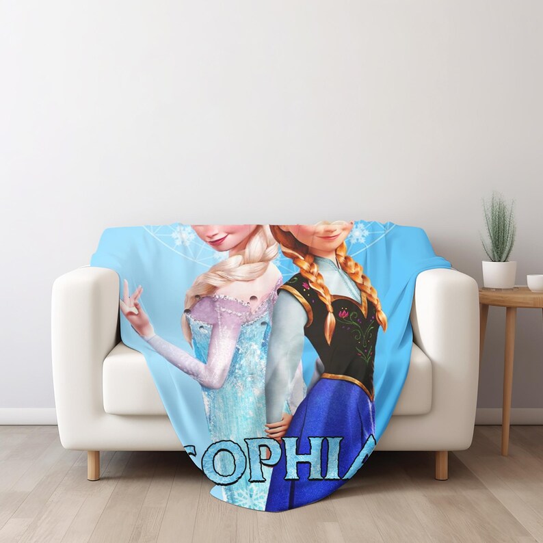 Może przedstawiać: Jasnoniebieski koc z Elsą i Anną z Krainy Lodu, z imieniem "SOPHIA" dużymi, niebieskimi literami. Elsa ma na sobie jasnoniebieską sukienkę, a Anna czarno-niebieską sukienkę. Koc jest narzucony na białą sofę.