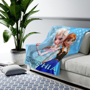 Może przedstawiać: Jasnoniebieski polarowy koc z Elsą i Anną z Krainy Lodu, z napisem "SOPHIA" dużymi niebieskimi literami. Koc jest narzucony na białą sofę, z zieloną rośliną w tle.