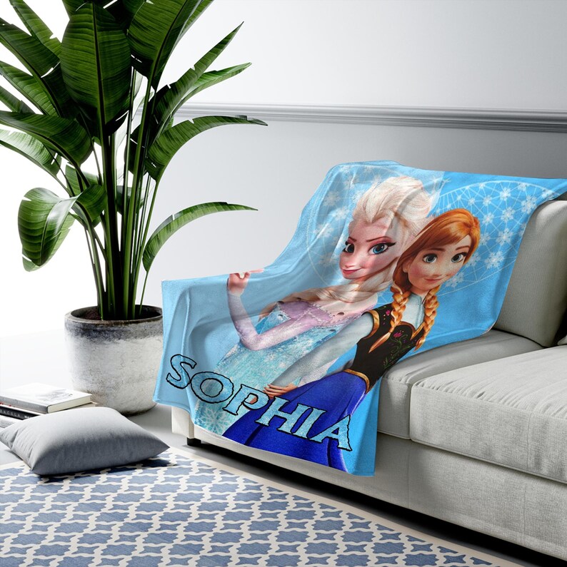 Może przedstawiać: Jasnoniebieski polarowy koc z Elsą i Anną z Krainy Lodu, z napisem "SOPHIA" dużymi niebieskimi literami. Koc jest narzucony na białą sofę, z zieloną rośliną w tle.