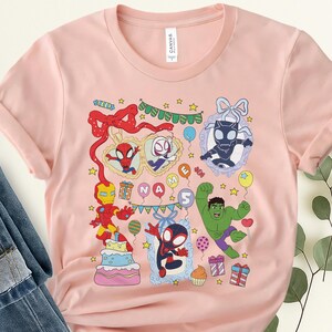 May include: A light pink short-sleeve t-shirt featuring a colorful graphic print of various superheroes, a birthday cake, presents, and balloons. The design includes the text "NAME" and "5" on balloons, surrounded by festive party decorations.
