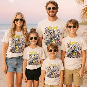 Pode incluir: Uma fam&iacute;lia de cinco pessoas vestindo camisetas creme combinando com um design gr&aacute;fico colorido com personagens, um bolo e o n&uacute;mero 3. As camisas t&ecirc;m a palavra "NAME" impressa acima do gr&aacute;fico.