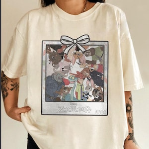 Puede incluir: Camiseta color crema con un dise&ntilde;o enmarcado de personajes de la pel&iacute;cula de Disney Zootopia. El dise&ntilde;o incluye un lazo blanco y las palabras "Zootopia 2026". La camiseta es una prenda casual para uso diario.
