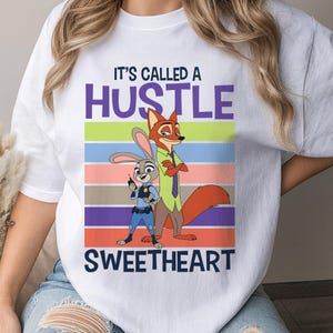May include: A white short-sleeved t-shirt featuring a graphic of Judy Hopps and Nick Wilde from Zootopia. The text reads IT'S CALLED A HUSTLE SWEETHEART above and below the characters, set against a background of horizontal colorful stripes.