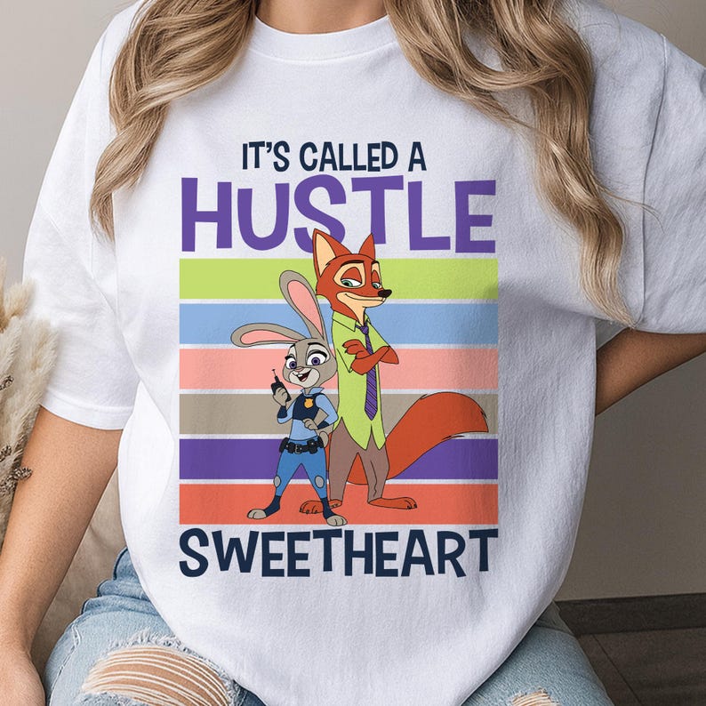 May include: A white short-sleeved t-shirt featuring a graphic of Judy Hopps and Nick Wilde from Zootopia. The text reads IT'S CALLED A HUSTLE SWEETHEART above and below the characters, set against a background of horizontal colorful stripes.
