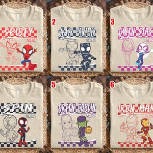 May include: Six beige t-shirts with superhero designs. Each shirt displays a different character's name and the year 2021. The designs include Spidey, Black Panther, Gwen Stacy, Spin, Green Goblin, and Iron Man.