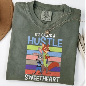 May include: An olive green crew neck t-shirt featuring a graphic of a cartoon rabbit and fox. The text reads IT'S CALLED A HUSTLE SWEETHEART above and below the characters, set against a background of horizontal stripes in blue, green, and pink.