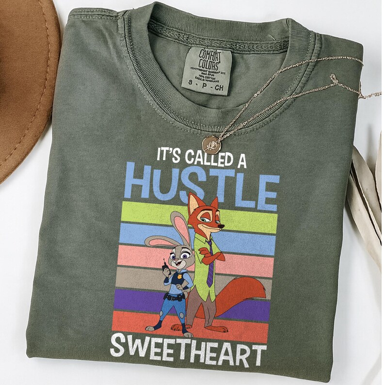 May include: An olive green crew neck t-shirt featuring a graphic of a cartoon rabbit and fox. The text reads IT'S CALLED A HUSTLE SWEETHEART above and below the characters, set against a background of horizontal stripes in blue, green, and pink.