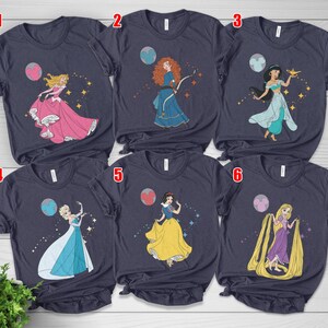 May include: Navy blue t-shirts featuring illustrations of Disney princesses. Each shirt showcases a different princess, including Aurora, Merida, Jasmine, Elsa, Snow White, and Rapunzel, with celestial and sparkling accents. A fun and colorful design for fans of Disney.