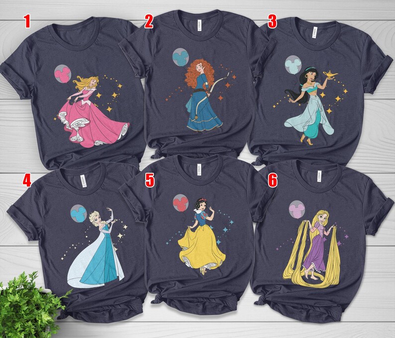May include: Navy blue t-shirts featuring illustrations of Disney princesses. Each shirt showcases a different princess, including Aurora, Merida, Jasmine, Elsa, Snow White, and Rapunzel, with celestial and sparkling accents. A fun and colorful design for fans of Disney.