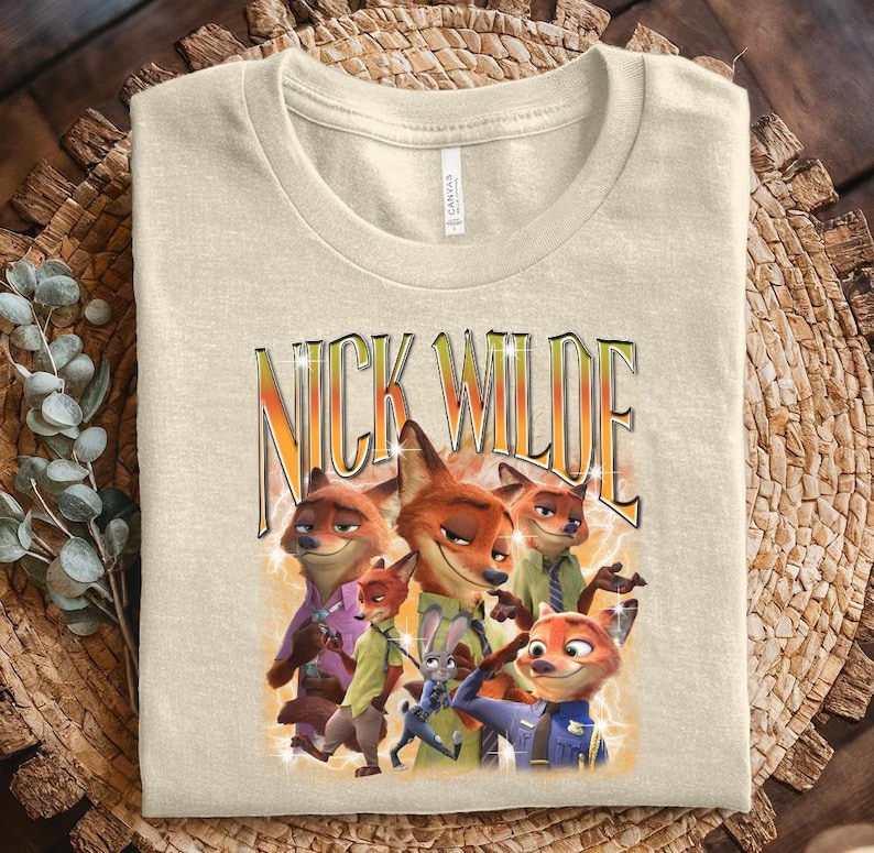 May include: Beige t-shirt featuring a graphic of Nick Wilde from Zootopia. The design includes the text "NICK WILDE" in a stylized font, with images of the character in various poses. The shirt is folded on a woven surface.
