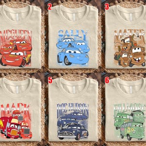 May include: Six beige t-shirts with different graphic designs featuring characters from the animated movie "Cars." The designs include "McQueen," "Sally," "Mater," "Mack," "Doc Hudson," and "Fillmore," each with corresponding car illustrations.