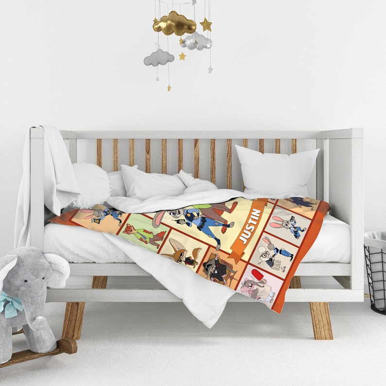 May include: A personalized baby blanket featuring cartoon characters in a grid pattern. The blanket is orange-bordered and has the name "Justin" printed on it. The blanket is draped over a white crib with a plush elephant toy in the foreground.