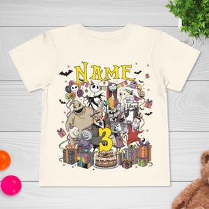 May include: Cream-colored t-shirt featuring a colorful graphic of characters from The Nightmare Before Christmas, with the word "NAME" above the characters and the number "3" below. The design includes bats, presents, and a birthday cake.