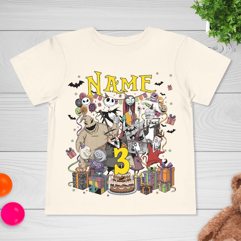 May include: Cream-colored t-shirt featuring a colorful graphic of characters from The Nightmare Before Christmas, with the word "NAME" above the characters and the number "3" below. The design includes bats, presents, and a birthday cake.