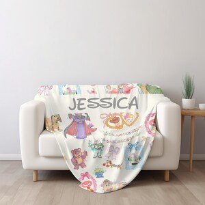 Puede incluir: Una manta crema con personajes de dibujos animados y el nombre "JESSICA" en letras grises. La manta est&aacute; sobre un sof&aacute; blanco. Una mesa auxiliar de madera y una planta est&aacute;n en el fondo.