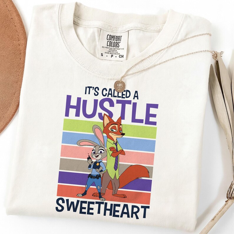 May include: A white short-sleeved t-shirt featuring a graphic of a rabbit and a fox. The text reads IT'S CALLED A HUSTLE SWEETHEART. The design includes horizontal stripes in green, blue, pink, and orange, with a Comfort Colors brand label at the neck.