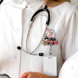 Op de afbeelding: Een intrekbare badge reel met cartoon auto's en Mickey Mouse-personages die hartvormige ballonnen in verschillende kleuren vasthouden. De badge reel is bevestigd aan een wit koord en wordt gedragen op een wit overhemd, met een stethoscoop op de achtergrond.