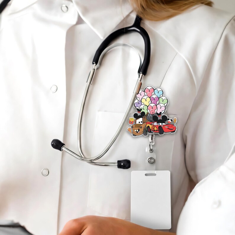 Op de afbeelding: Een intrekbare badge reel met cartoon auto's en Mickey Mouse-personages die hartvormige ballonnen in verschillende kleuren vasthouden. De badge reel is bevestigd aan een wit koord en wordt gedragen op een wit overhemd, met een stethoscoop op de achtergrond.