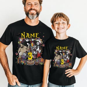 Pode incluir: T-shirts pretas com um gr&aacute;fico colorido com personagens de "O Estranho Mundo de Jack" e o n&uacute;mero "3". As camisas t&ecirc;m a palavra "Name" acima do gr&aacute;fico e s&atilde;o usadas por um homem e uma crian&ccedil;a.