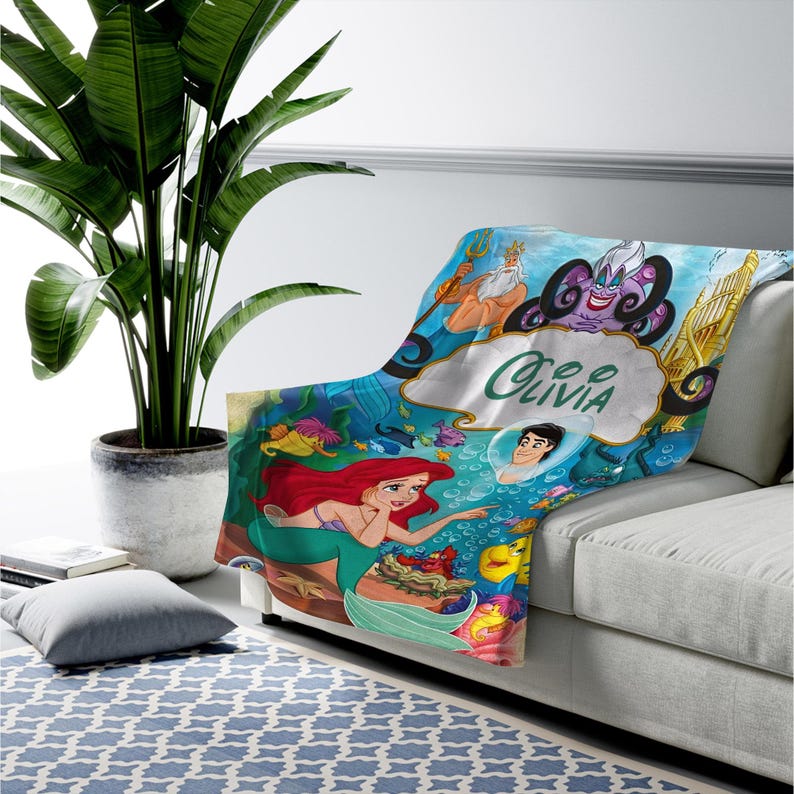 May include: A soft throw blanket featuring a vibrant underwater scene with characters from The Little Mermaid, including Ariel, King Triton, Ursula, and Prince Eric. The center displays a white cloud-shaped graphic with the name Olivia printed.