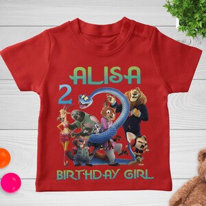 May include: Red short-sleeved t-shirt featuring a colorful graphic of animated characters and the text "ALISA 2 BIRTHDAY GIRL". The design includes a blue snake and various animal figures.