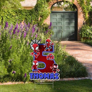 May include: A red and blue yard sign in the shape of the number 5, featuring Spider-Man characters. The sign has the name "THOMAS" in blue letters at the bottom. The sign is placed in a grassy yard with a garden in the background.