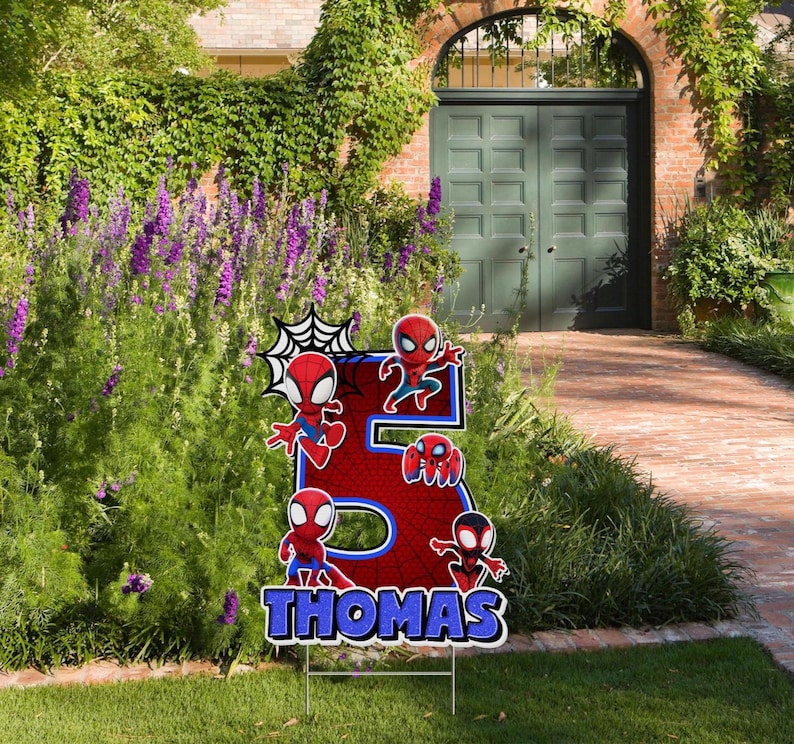 May include: A red and blue yard sign in the shape of the number 5, featuring Spider-Man characters. The sign has the name "THOMAS" in blue letters at the bottom. The sign is placed in a grassy yard with a garden in the background.