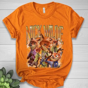 May include: Orange t-shirt featuring a graphic of Nick Wilde from Zootopia. The design includes multiple images of the character with the text "NICK WILDE" in gold lettering. The shirt is made of a soft material.
