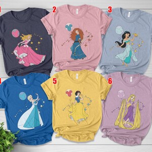 May include: Six short-sleeved t-shirts in various colors, each featuring a different princess graphic. The designs include Aurora, Merida, Jasmine, Elsa, Snow White, and Rapunzel, with accompanying star and balloon accents. The shirts are displayed on a light wood surface.