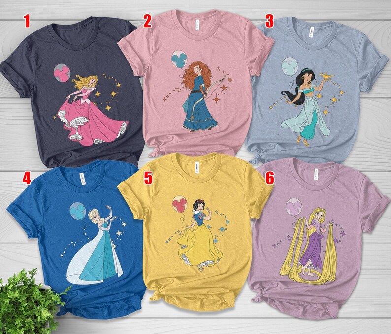 May include: Six short-sleeved t-shirts in various colors, each featuring a different princess graphic. The designs include Aurora, Merida, Jasmine, Elsa, Snow White, and Rapunzel, with accompanying star and balloon accents. The shirts are displayed on a light wood surface.