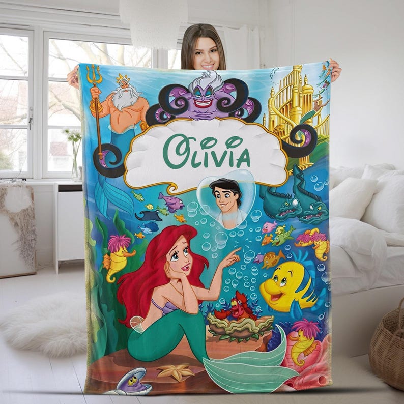 May include: A rectangular fleece blanket featuring a colorful underwater scene with characters from The Little Mermaid, including Ariel, Flounder, King Triton, and Ursula. The center displays a white shell shape with the name Olivia printed in green.