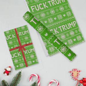 May include: Green wrapping paper with a white snowflake pattern and the words "FUCK TRUMP" printed on it. A wrapped gift with a red ribbon and the words "FUCK TRUMP" is also visible. Festive holiday decor is scattered around the wrapping paper.