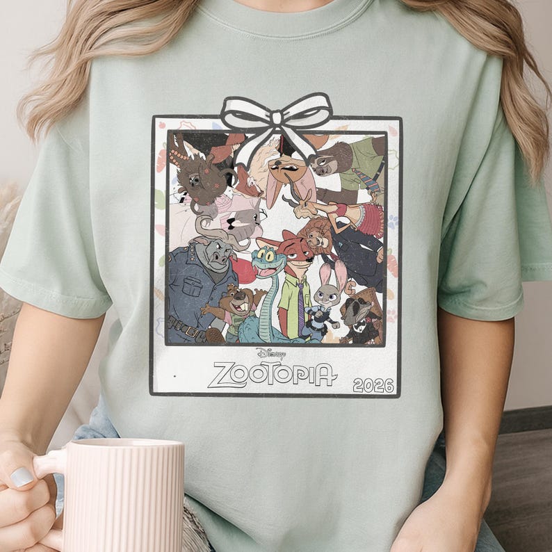 Puede incluir: Camiseta verde claro con un gr&aacute;fico enmarcado de personajes de la pel&iacute;cula de Disney Zootopia. El gr&aacute;fico incluye un lazo blanco en la parte superior y el t&iacute;tulo de la pel&iacute;cula y el a&ntilde;o 2026 en la parte inferior.