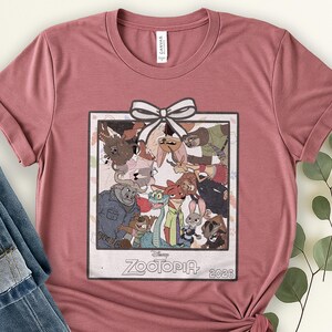 Puede incluir: Camiseta rosa polvoriento con un gr&aacute;fico de la pel&iacute;cula de Disney Zootopia. El gr&aacute;fico es una imagen enmarcada de los personajes con un lazo blanco en la parte superior. El texto "Zootopia" y "2026" est&aacute; en la parte inferior de la imagen.