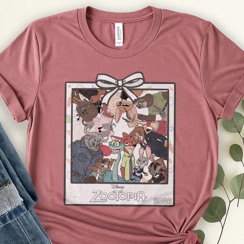 Puede incluir: Camiseta rosa polvoriento con un gr&aacute;fico de la pel&iacute;cula de Disney Zootopia. El gr&aacute;fico es una imagen enmarcada de los personajes con un lazo blanco en la parte superior. El texto "Zootopia" y "2026" est&aacute; en la parte inferior de la imagen.