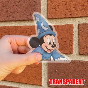 Puede incluir: Una pegatina transparente con Mickey Mouse vestido con un atuendo de mago azul, con una luna y estrellas en su sombrero. La pegatina tiene la palabra "TRANSPARENT" en letras rojas.