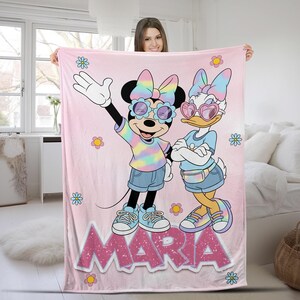 Op de afbeelding: Een roze deken met Minnie Mouse en Daisy Duck in zonnebrillen en tie-dye outfits. De deken heeft de naam "MARIA" in grote, glinsterende letters en is versierd met bloemen.