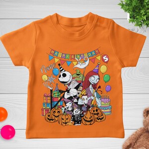 May include: Orange short-sleeved t-shirt featuring a colorful graphic of characters from The Nightmare Before Christmas. The design includes Jack Skellington, Sally, and pumpkins, with a "Happy Birthday" banner and a birthday cake.