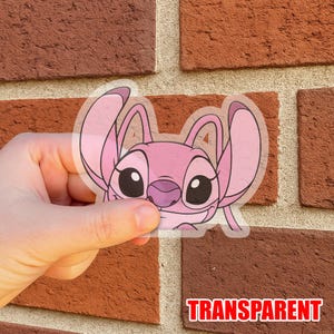 May include: A transparent sticker featuring a pink cartoon character with large eyes and ears. The sticker has the word "TRANSPARENT" in a red font at the bottom. The sticker is held in front of a brick wall.