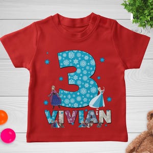 May include: Red toddler t-shirt featuring a large blue number 3 filled with white snowflakes. The name "VIVIAN" is printed below the number. The shirt has images of animated characters and blue snowflake accents.